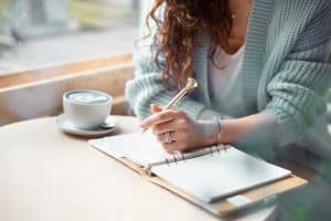 Young woman in blue sweater sitting by a window and journaling